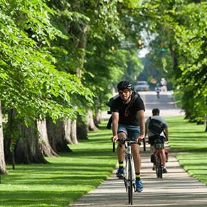 Student on bike on sidewalk on campus with green grass on either side. 